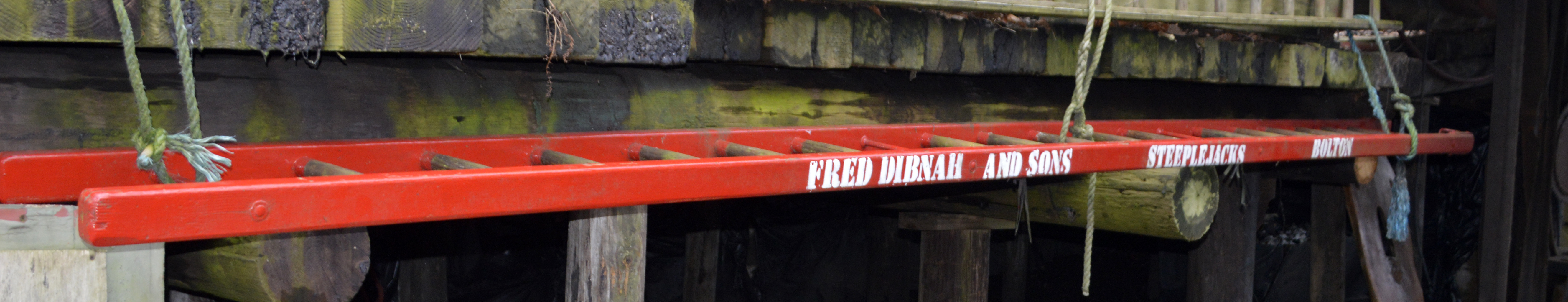 An iconic red painted wooden steeplejack ladder bearing the name 'Fred Dibnah and Sons Steeplejacks Bolton'.